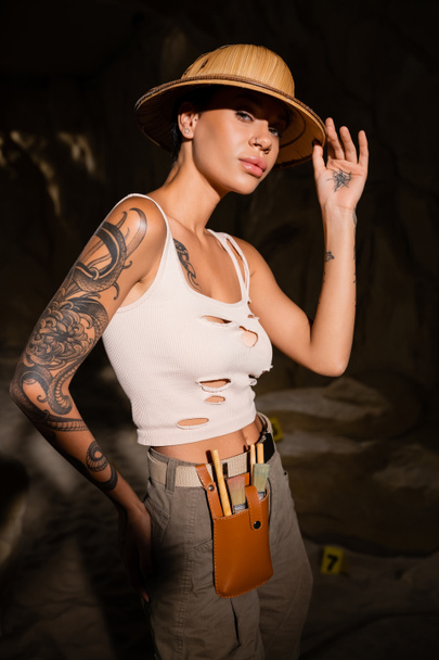 sexy tatuaż archeolog w zbiornik top dotykając safari kapelusz i patrząc w aparat fotograficzny w ciemnej jaskini - Zdjęcie, obraz