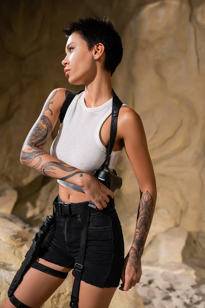 sexy archaeologist in white crop top and black shorts taking gun out of holster in cave - Foto, afbeelding