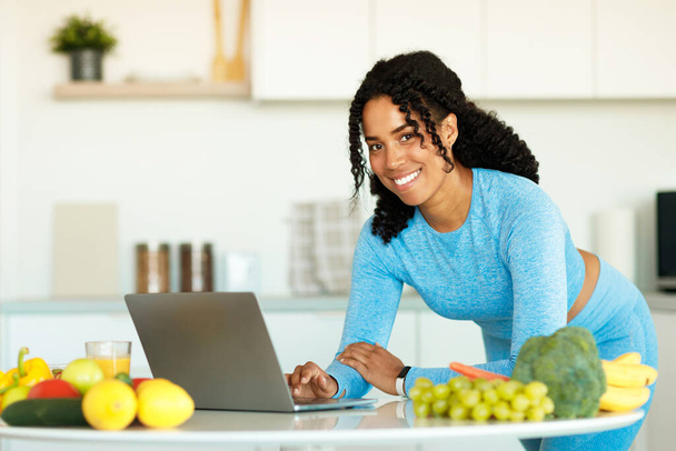 Healthy lifestyle concept. Happy african american woman using laptop and searching new recipe for lunch, preparing fresh vegetable salad in kitchen, free space - Foto, immagini