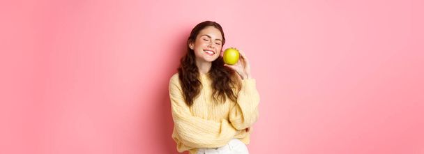 Healthcare, stomatology and people concept. Happy girl show her white perfect teeth, smile and green apple, eating healthy food and fruits, standing against pink background. - Photo, Image