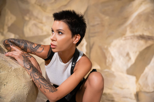 brunette tatuoitu arkeologi katselee pois piiloutuessaan kiven takana autiomaassa - Valokuva, kuva