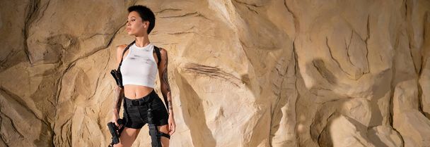 getatoeëerde jonge archeoloog in sexy outfit met pistool en staan in de buurt van rotsen, banner - Foto, afbeelding