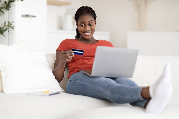 Sonriente mujer afroamericana haciendo pagos en línea con computadora portátil y tarjeta de crédito mientras está sentada en el sofá en casa, feliz mujer negra comprando en Internet mientras descansa con la computadora en la sala de estar - Foto, Imagen