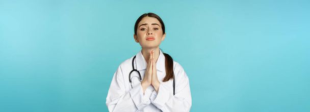 Vrouwelijke arts bedelen, smeken of smeken, doen wens, vragen om hulp, staan wanhopig tegen torquoise achtergrond. - Foto, afbeelding