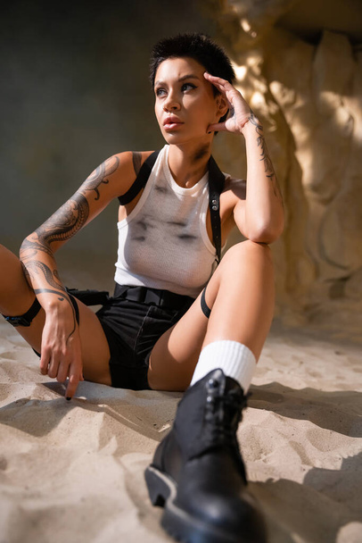 sexy young archaeologist with tattoo and short hair sitting in dirty clothes on sand - Foto, immagini