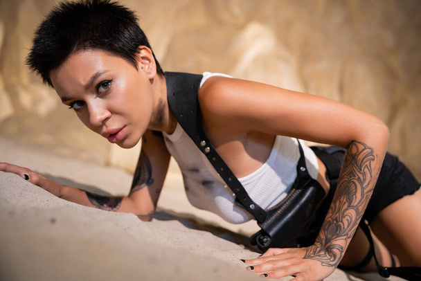 sexy and tattooed archaeologist in dirty clothes and holster crawling on sand in cave - Foto, Imagem