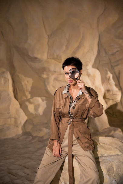 young archaeologist with tattoo holding magnifying glass near face in cave - Foto, Imagen