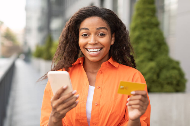 Satisfecho inspirado sorprendió a la mujer afroamericana milenaria con tarjeta de crédito y uso de teléfono inteligente para pagar dinero en la calle de la ciudad. Nueva aplicación para compras en línea, la venta de reembolso y ganar, grandes noticias y anuncios - Foto, imagen