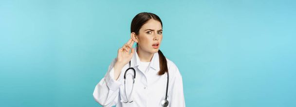 Bild einer verwirrten Ärztin, die dich nicht hören kann, die Hand am Ohr hält und verwirrt guckt, lauter spricht, torquoise Hintergrund. - Foto, Bild