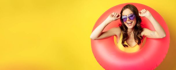 Close up portrait of stylish young woman, girl with swimming ring and summer sunglasses, ready for beach holiday, vacation, standing over yellow background. - Foto, Imagen