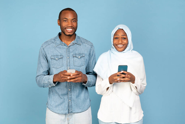 Afrikkalainen amerikkalainen islamilainen pari Kännyköiden avulla Tekstiviesti seisoo Blue Studio tausta, hymyilee kameralle. Puolisot käyttävät uutta sovellusta ja selailu Internet Via älypuhelimet - Valokuva, kuva