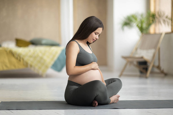 Evde oturma odasında antrenman yaparken, yoga minderinde bebek bekleyen ve göbeğini kucaklayan aktif giyimde gülümseyen hamile kadın meditasyon yapıyor, Uzay 'ı kopyalıyor. - Fotoğraf, Görsel