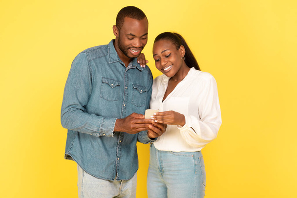 Веселые афроамериканские супруги используют смартфон вместе, общаясь онлайн, стоя в студии на жёлтом фоне. Пара с использованием нового мобильного приложения. Технологии и гаджеты - Фото, изображение