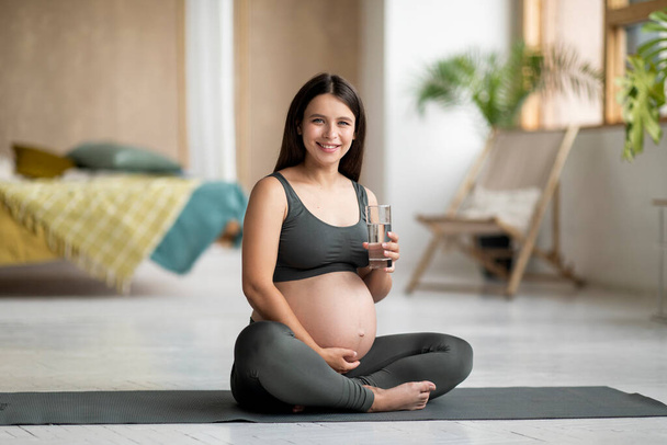 Terveellistä elämäntapaa. Nuori raskaana oleva nainen istuu joogamatolla ja pitää lasia vedellä, kaunis odottava nainen nauttii virkistävästä juomasta rentoutuessaan meditaation jälkeen kotona, kopioi tilaa - Valokuva, kuva