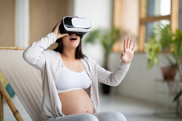 Young Pregnant Woman Using VR Glasses While Relaxing In Chair At Home, Excited Expectant Female With Big Belly Experiencing Virtual Reality, Enjoying Modern Technologies For Leisure, Free Space - Foto, Imagen