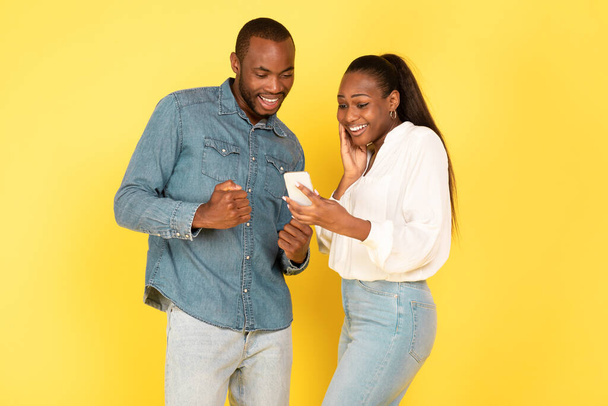 Joyful Black Spouses Using Smartphone And Shaking Fists In Joy Standing Over Yellow Studio Background. Couple Reading Great News. Wow Application Or Offer Advertisement - Photo, Image