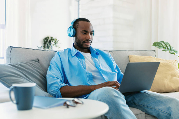 Czarny człowiek w bezprzewodowych słuchawkach, korzystający z laptopa, siedzący na kanapie w domu, pracujący online w domu. Afrykański amerykański facet za pomocą komputera i surfing internet - Zdjęcie, obraz