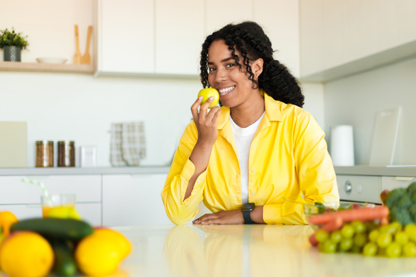 Felice donna nera attraente mangiare frutta, seduto sul tavolo in cucina e sorridente alla macchina fotografica, spazio libero. Bella signora godendo di mangiare mela, cibi sani per la salute benessere per perdere peso - Foto, immagini