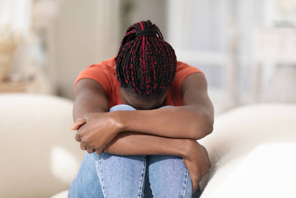 Desglose mental. Retrato de una joven afroamericana trastornada llorando en casa, una mujer negra deprimida enterrando la cabeza en las rodillas mientras está sentada en el sofá, sintiéndose sola y triste, copiando el espacio - Foto, imagen
