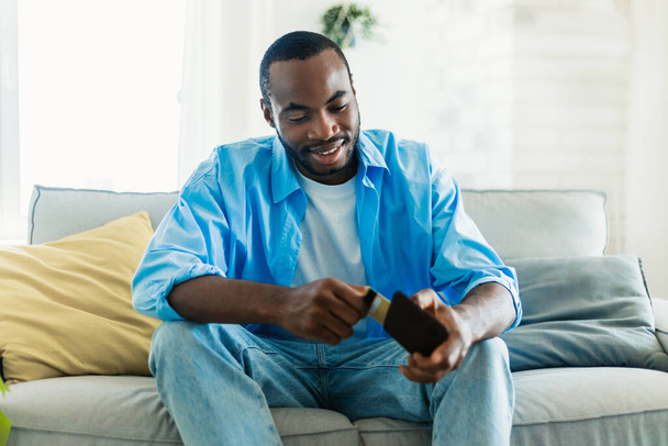 Feliz hombre afroamericano sonriendo mientras toma la tarjeta de crédito de la cartera, sentado en el sofá en el interior de la sala de estar, espacio de copia gratuita. Concepto de dinero electrónico - Foto, Imagen