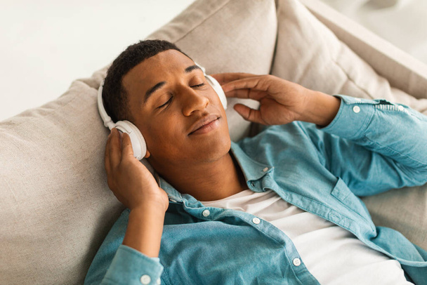 Satisfied young black male in jeans and wireless headphones with closed eyes lies on couch, enjoys favorite music, comfort and lazy at free time alone in room. Rest and relax at home with audio app - Фото, изображение