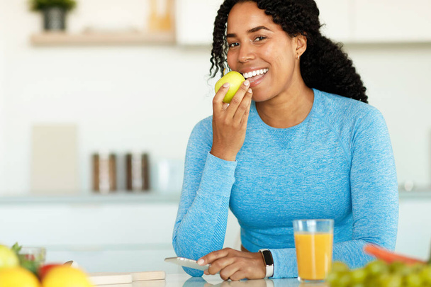 Active african american woman biting apple and looking at camera, sitting in kitchen interior and having lunch after workout at home. Sport lady enjoy eating fruits for health after exercise - Φωτογραφία, εικόνα