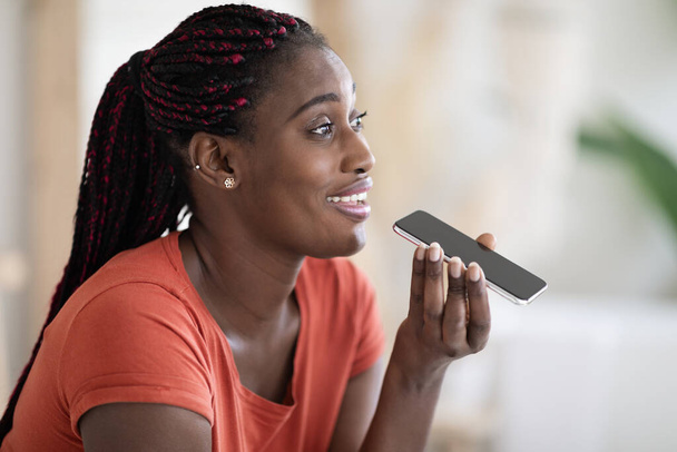 Primo piano girato di felice nero donna registrazione messaggio vocale su smartphone mentre si rilassa a casa, sorridente afro-americano femminile utilizzando assistente virtuale sul telefono cellulare, godendo di tecnologie moderne - Foto, immagini