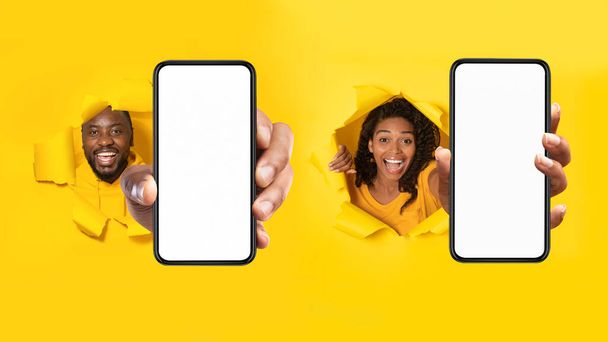 Black Man And Woman Showing Phones With Blank Screens Stretching Arm To Camera Through Hole In Torn Yellow Paper Background. Mobile Application Advertisement. Panorama, Mockup - Photo, Image