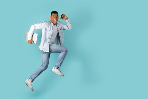 Χαμογελώντας ενθουσιασμένοι χιλιετή αφροαμερικανός τύπος με ανοιχτό στόμα με φορητό υπολογιστή άλμα στον αέρα στο κενό χώρο, απομονώνονται σε μπλε φόντο, στούντιο. Ανθρώπινα συναισθήματα, τεχνολογία για την εργασία, μεγάλη προσφορά και διαφήμιση - Φωτογραφία, εικόνα
