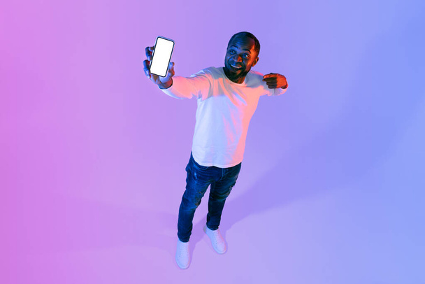 Cheerful attractive stylish african american mature man showing cell phone with white empty screen, recommending nice offer or mobile app, mockup, neon studio background, high angle shot, copy space - Foto, afbeelding