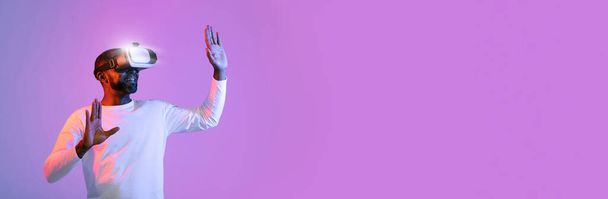 Excited middle aged black man in white using VR glasses on neon studio background, touching something invisible with hands, panorama, copy space. Modern technologies, entertainment, virtual reality - Фото, зображення