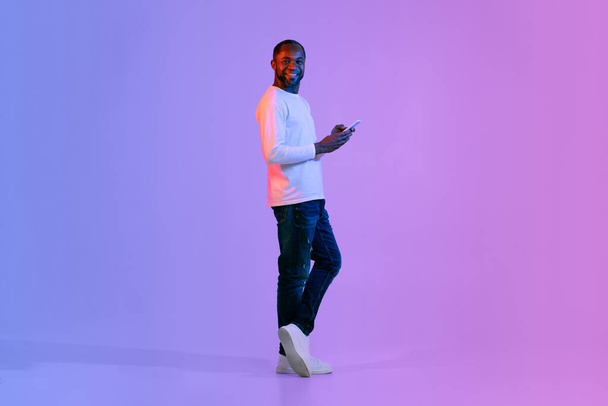 Vrolijke knappe zwarte man van middelbare leeftijd in stijlvolle casual outfit met behulp van moderne smartphone en glimlachen over neon studio achtergrond, gokken op internet, chatten met dames, kopieerruimte, volledige lengte - Foto, afbeelding