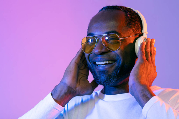 Retrato de primer plano de un hombre negro de mediana edad feliz y guapo escuchando música y sonriendo sobre el fondo de neón, usando auriculares inalámbricos modernos, usando anteojos amarillos, mirando el espacio de copia - Foto, Imagen