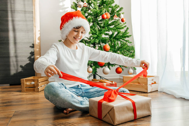 A happy girl in a santa claus hat opens a Christmas present, unties the ribbon on the package. Christmas and New Year. - Photo, Image