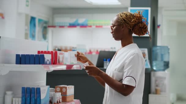 Glimlachende Afro-Amerikaanse apotheker het controleren van drugs op rekken in de apotheek, het helpen van klanten met medicijnen, het houden van pillen flessen en dozen vitaminen. Werken bij de apotheek. Handschot. - Video