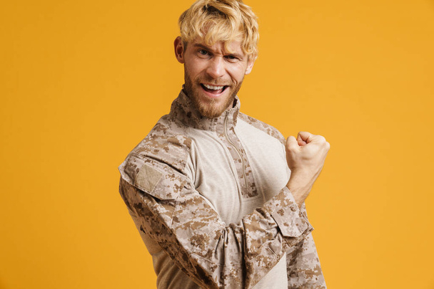European military man wearing uniform showing his bicep at camera isolated over yellow background - Фото, зображення