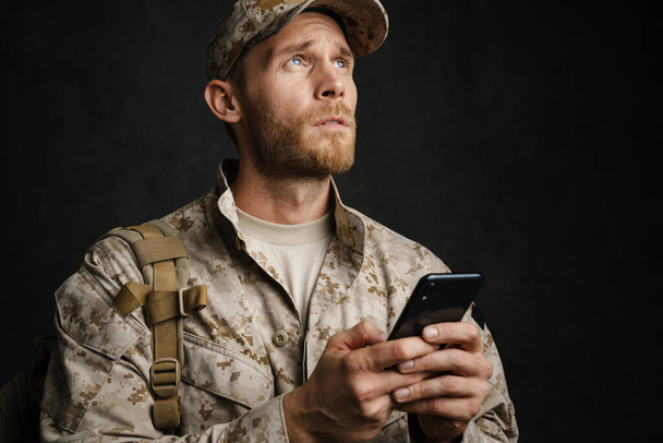 European military man wearing uniform using mobile phone isolated over black background - Photo, Image