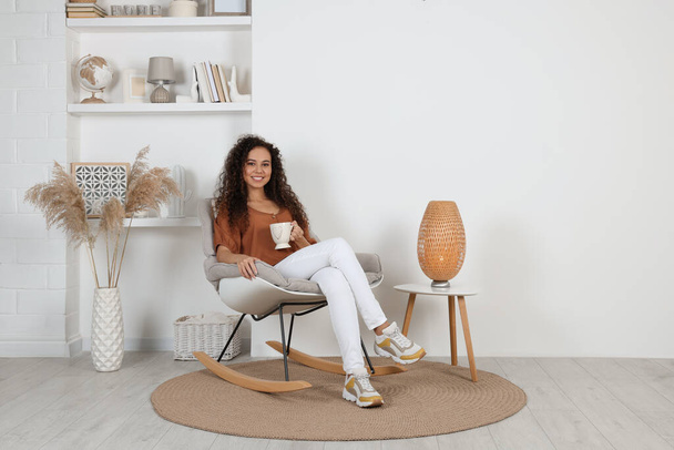 Young African-American woman with cup of drink sitting in rocking chair at home - Photo, image