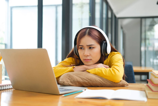 Young female student or worker using laptop to study or work online and feeling bored, tired, and disappointed while sitting and leaning on her desk. Online learning or work concept. - Photo, Image