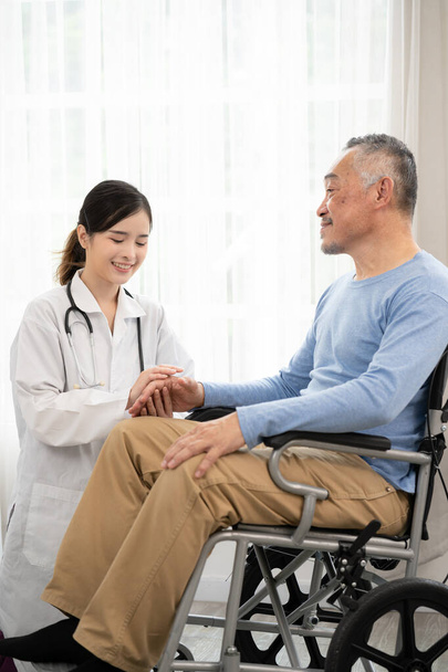 年配の男が車椅子に腰掛けている間手をつないでいるアジア系女性医師。車椅子の幸せな高齢者が彼女と手を取り合いながら医者とコミュニケーションをとる. - 写真・画像