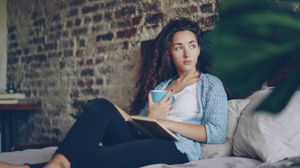 Cheerful girl is reading funny book in bedroom lying on comfy double bed, smiling and holding cup with drink. Hobby, happy people, beautiful interiors and literature concept. - Фото, изображение