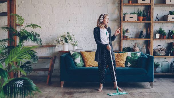 Beautiful young lady is listening to music through wireless headphones and doing housework cleaning the floor with mop. Girl is dancing and singing enjoying cleanup at home. - Foto, Bild