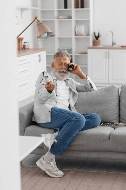 Senior bearded man talking by mobile phone on sofa in kitchen - Valokuva, kuva