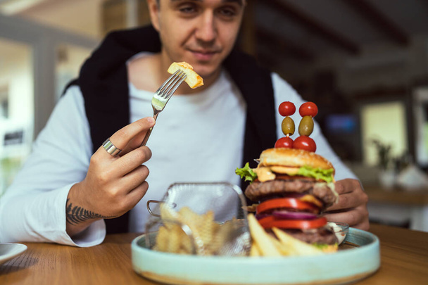 Один молодой взрослый белый мужчина сидит за столом в ресторане с французскими и бургерскими чипсами, которые едят в одиночестве в течение дня реальные люди копируют космический фокус на руку и вилку - Фото, изображение