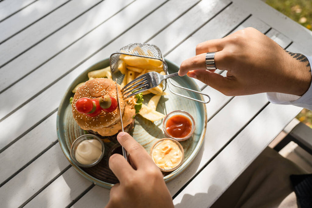 primer plano en la hamburguesa en un plato y las manos de hombre caucásico desconocido se preparan para comer en la mesa en el día soleado al aire libre en el restaurante mantenga tenedor y cuchillo - Foto, Imagen