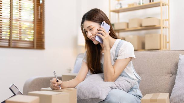 E-commerce businessman is talking on the phone with a customer who comes in to inquire and place her order, Woman writes mailbox address in preparation for delivery, SME entrepreneur, Packing box. - Zdjęcie, obraz