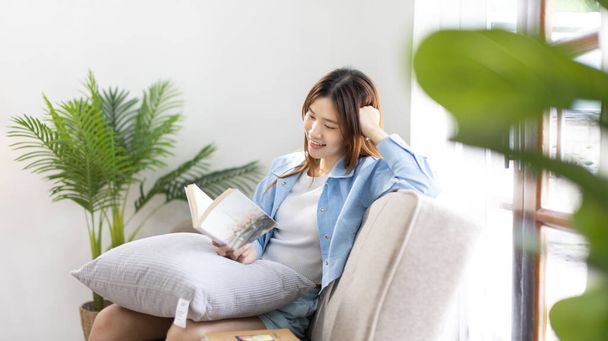 Asian woman reading a novel on the sofa in the living room,  Weekend leisure activities, happiness during vacation at home, Favorite corner of the house, Beautiful woman smiling sweetly. - Φωτογραφία, εικόνα