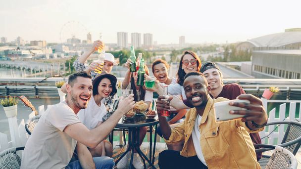 Young African American man is holding smartphone and taking selfie with his friends multi-ethnic group holding bottles and glasses, posing and enjoying rooftop party. - Fotografie, Obrázek