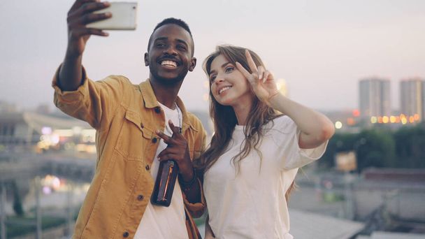 Attractive multiracial couple is taking selfie with smartphone at rooftop party, man and woman are holding drinks and posing with funny faces with evening city in background. - Zdjęcie, obraz