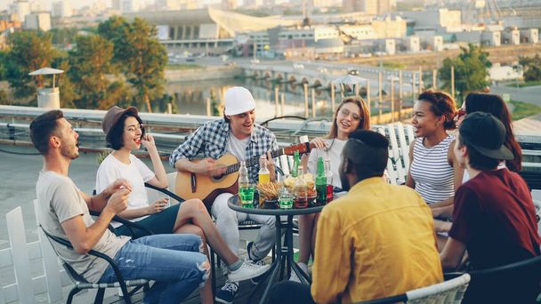Students are having fun on rooftop playing the guitar, singing and dancing sitting at table on rooftop enjoying free time. Modern lifestyle, joy and musical instruments concept. - Photo, Image
